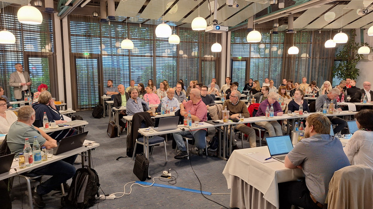 Delegierte im Saal mit Tischen in Reihenbestuhlung; vorne mit Blick zu den Delegierten die Bezirksleitung mit Beamer-Präsentation